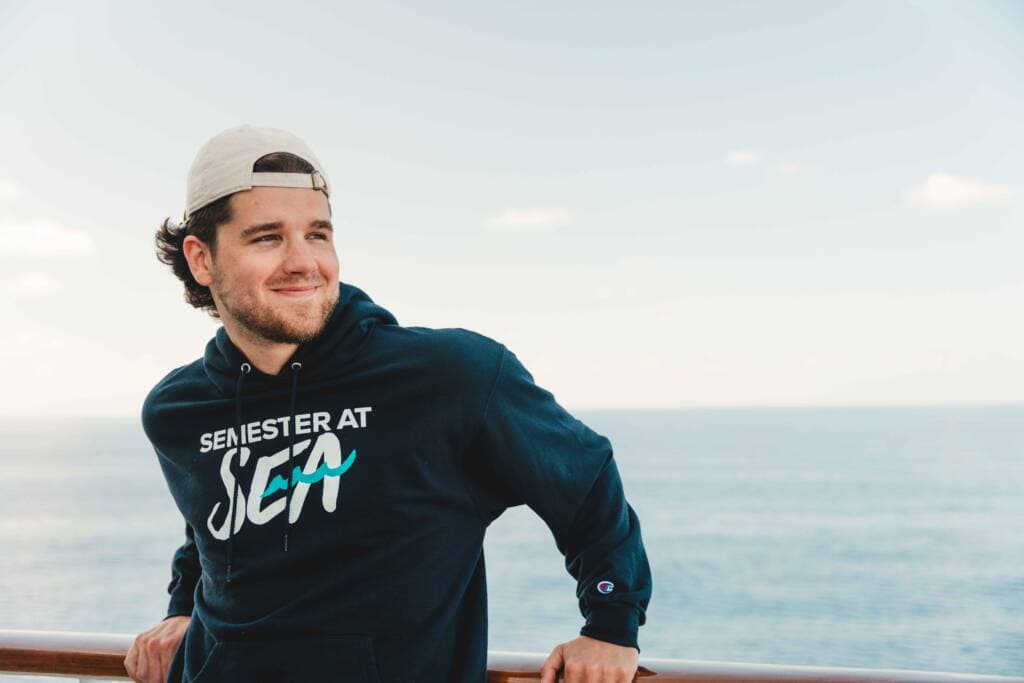 A young man in a dark blue hoodie and a backwards baseball cap leans against the railing of a ship and smiles at something out of frame. 