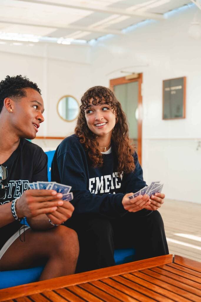 A young man and woman sit side-by-side, each holding playing cards and looking at one another knowingly. 