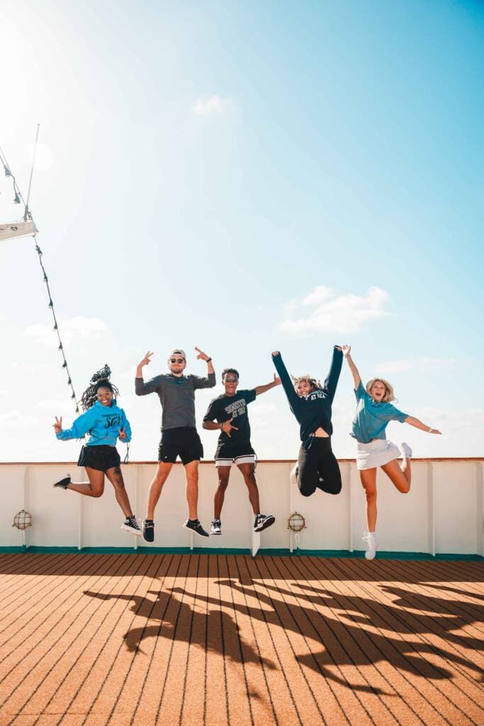 Five people jump into the air in unison, the bright sun casting shadows on the wooden deck beneath them. 